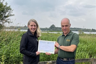Two people holding a certificate stood on grass at a wetland nature reserve