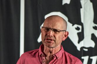 Man stood reading from book with black and white logo behind