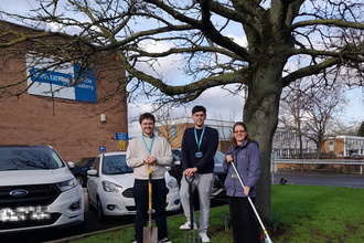 Three people infront of a tree holding gardening tools next to a car park and office building