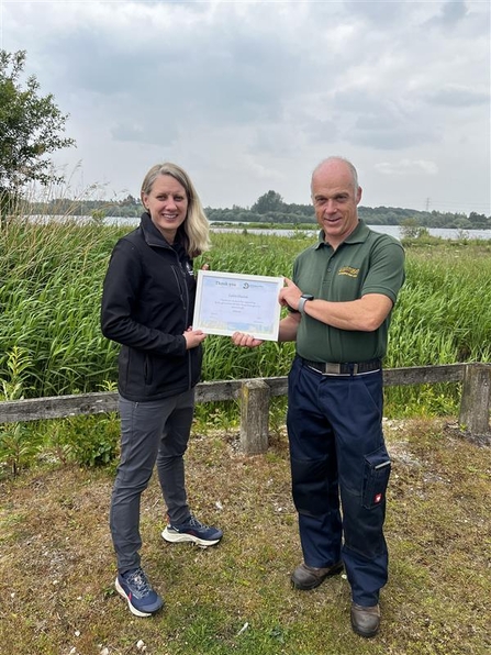 Two people holding a certificate stood on grass at a wetland nature reserve