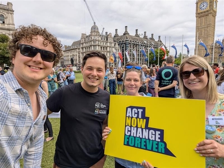 Four people took in front of Westminster, smiling broadly at the camera. They are holding a sign that says Act Now Change Forever. Behind them can be seen a crowd of people at the Mass Lobby