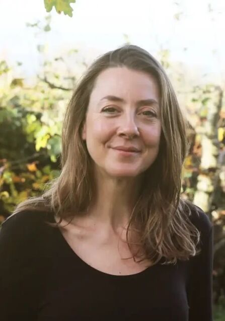 Woman with brown hair smiling in front of trees and hedges