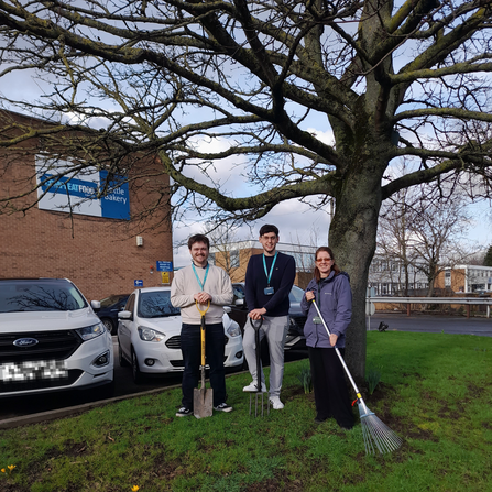 Three people infront of a tree holding gardening tools next to a car park and office building