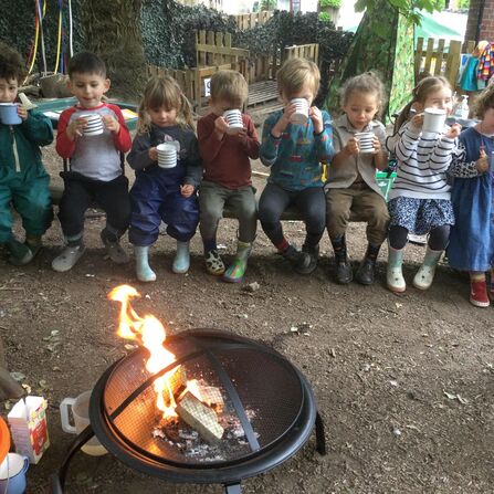 Children sat on a bench in a row infront of a campfire