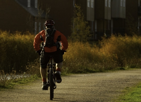 Man riding bike down path by a river with houses in background, The National Forest, UK 